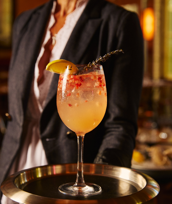 A peach-colored cocktail garnished with a lemon slice and herbs in a wine glass, held on a tray by a server in a blazer.