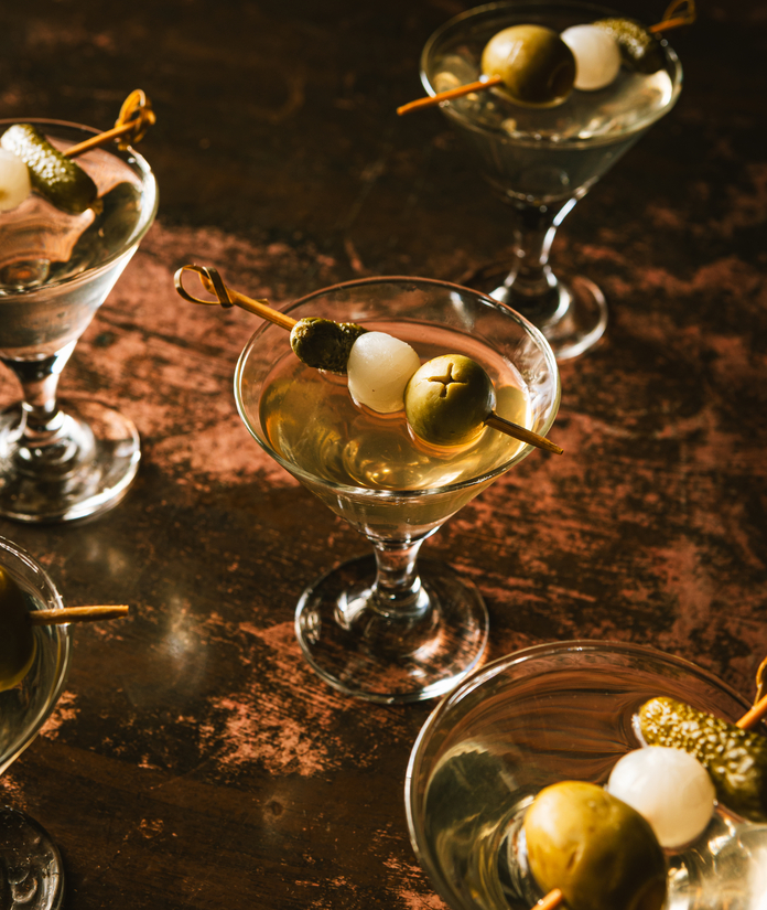 Several dirty martinis garnished with olives and cocktail onions on skewers, arranged on a dark surface.