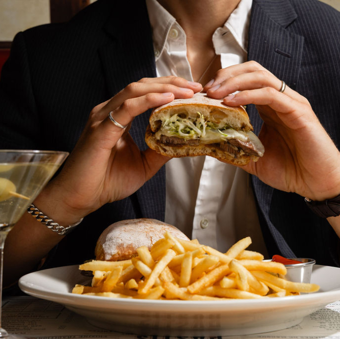 Person in business attire holding a burger with lettuce and meat, with a plate of french fries on the table