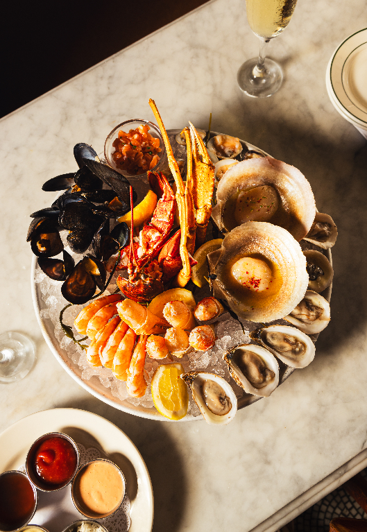 An overhead view of a seafood tower on ice with oysters, shrimp, mussels, lobster, and clams, served with dipping sauces and white wine.