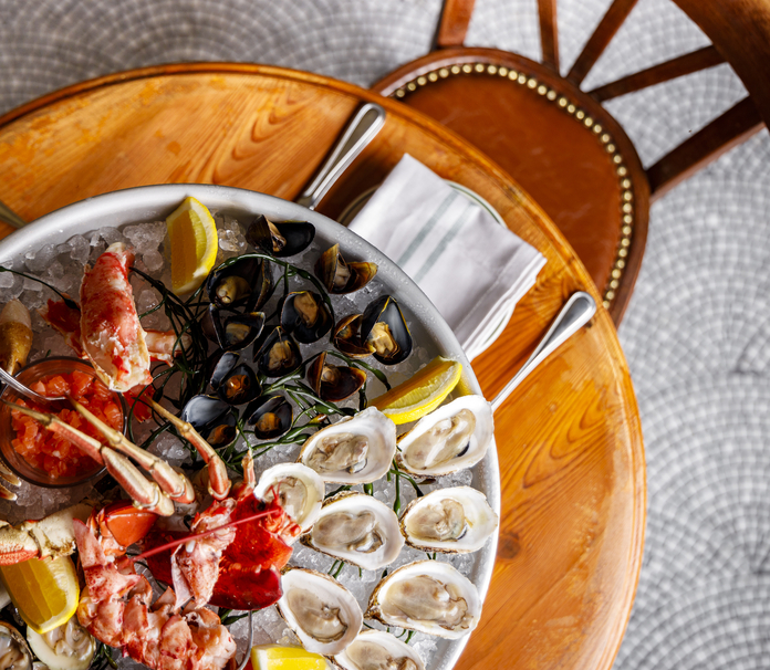 A wooden table set with various dishes, including roasted chicken, oysters on ice, and pasta, alongside glasses of white wine and menus.