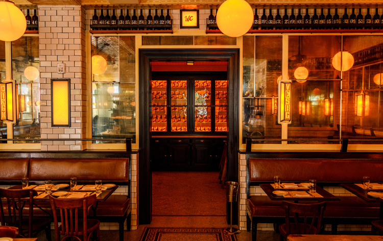 Cozy, vintage-style restaurant with wood tables, leather booth seating, hanging pendant lights, and framed artwork on white brick walls.
